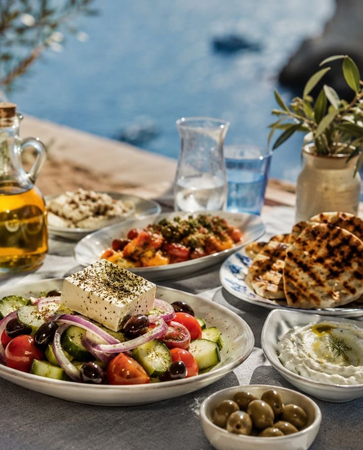A beautifully styled Greek food spread by the seaside featuring fresh Greek salad with feta cheese, olives, grilled pita bread, creamy tzatziki, and Mediterranean dishes on a rustic outdoor table under warm sunlight.