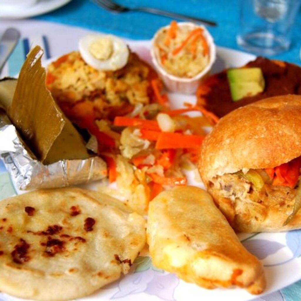 Top view of traditional Salvadoran food ingredients including corn, beans, rice, cheese, plantains, yuca, avocado, and pupusas arranged on a rustic table