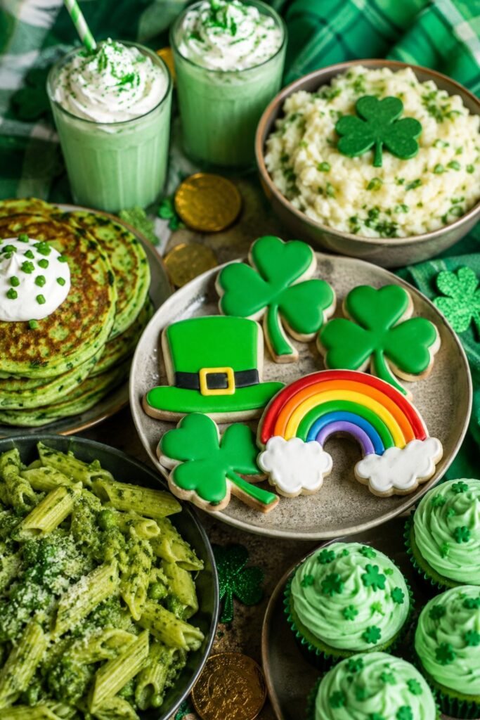 A vibrant St. Patrick’s Day food spread featuring green shamrock cookies, festive cupcakes, creamy mashed potatoes, green pancakes, pesto pasta, and mint-flavored drinks arranged on a rustic table with holiday decorations.